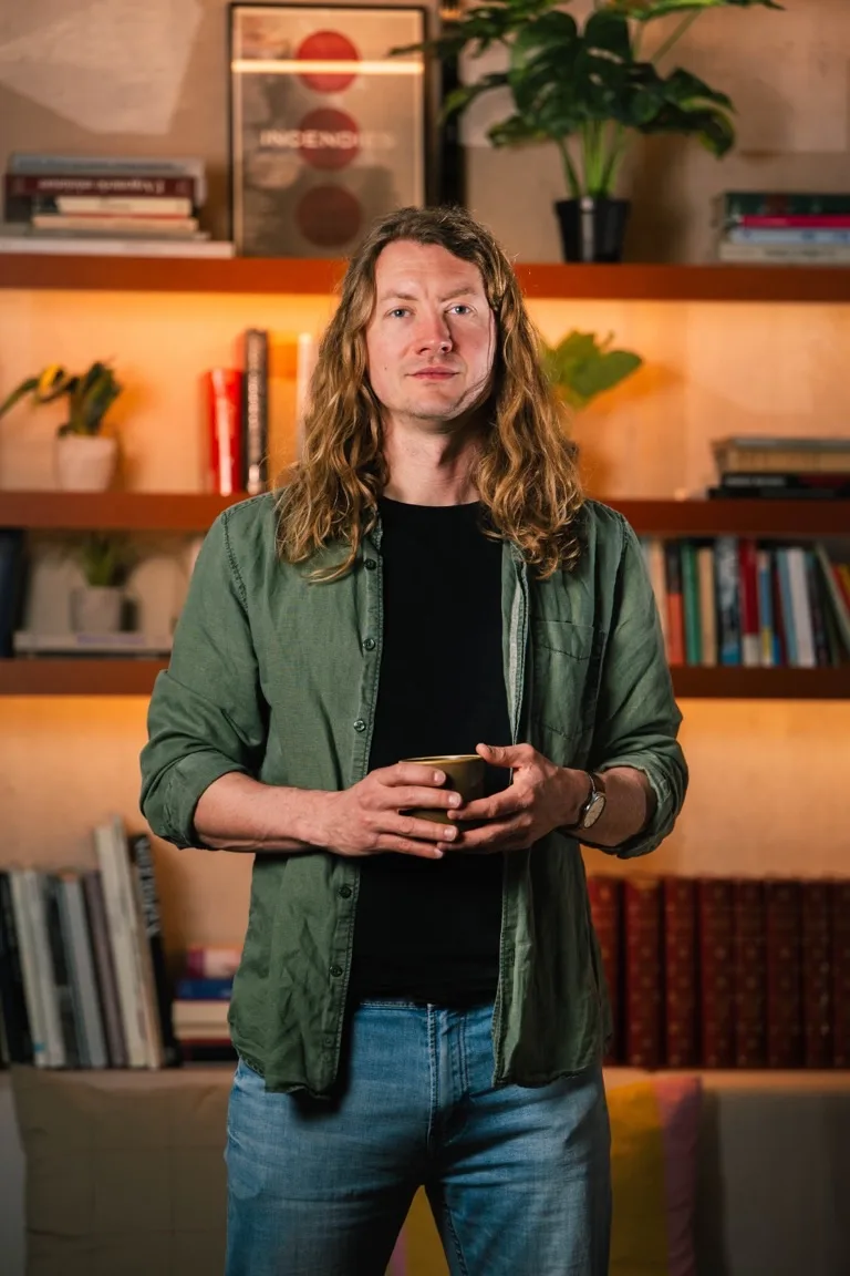 Portrait of a smiling man with long hair, standing in front of a bookshelf